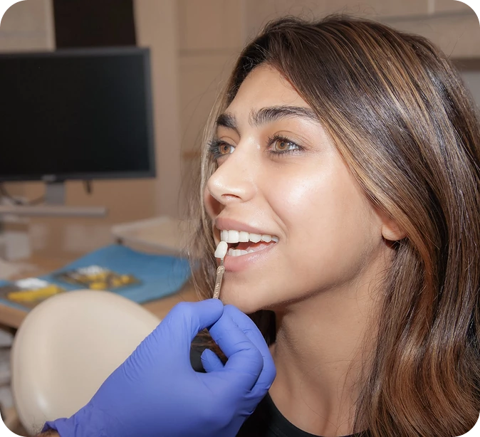 Dentist holding a shade guide next to a female patient’s teeth to match color for veneers or crowns.