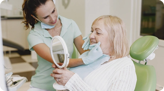 Female dentist holding a mirror for an elderly woman to check her smile after a dental treatment.