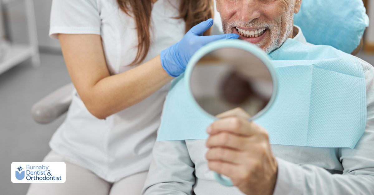 Senior patient sitting in a dental chair smiling while holding a hand mirror, reviewing the results of modern dental implant treatment